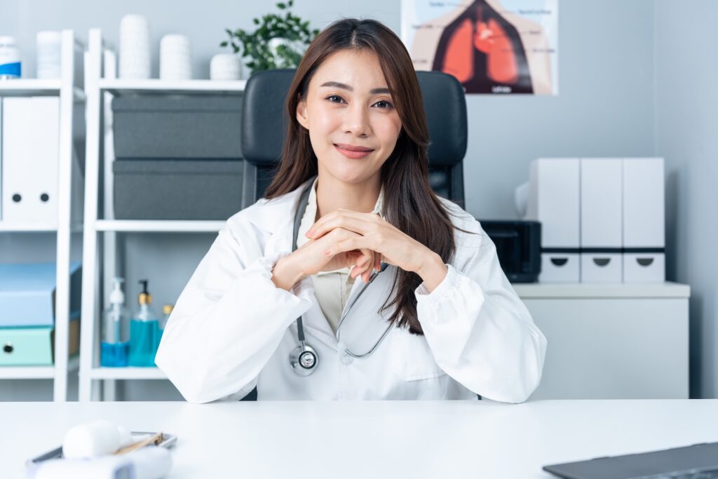 Photo of a doctor sitting at a desk with her hands clasped and her elbows on the desk. She is smiling.